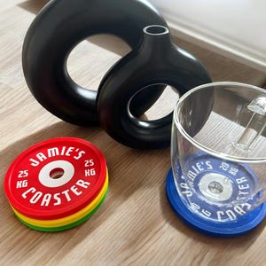 Puede incluir: Dos jarrones negros mate en forma de rosquilla, una pila de tres posavasos circulares en rojo, amarillo y verde con el texto JAMIE'S COASTER 25 KG, y una taza de cristal transparente sobre un posavasos azul con el texto JAMIE'S COASTER 20 KG.