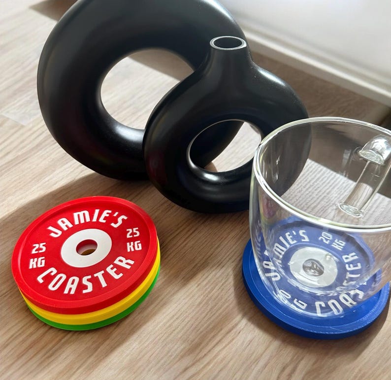 Puede incluir: Dos jarrones negros mate en forma de rosquilla, una pila de tres posavasos circulares en rojo, amarillo y verde con el texto JAMIE'S COASTER 25 KG, y una taza de cristal transparente sobre un posavasos azul con el texto JAMIE'S COASTER 20 KG.