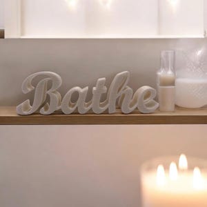 May include: White wooden sign with the word "Bathe" in a cursive font. The sign is sitting on a wooden shelf with two clear glass bottles behind it.
