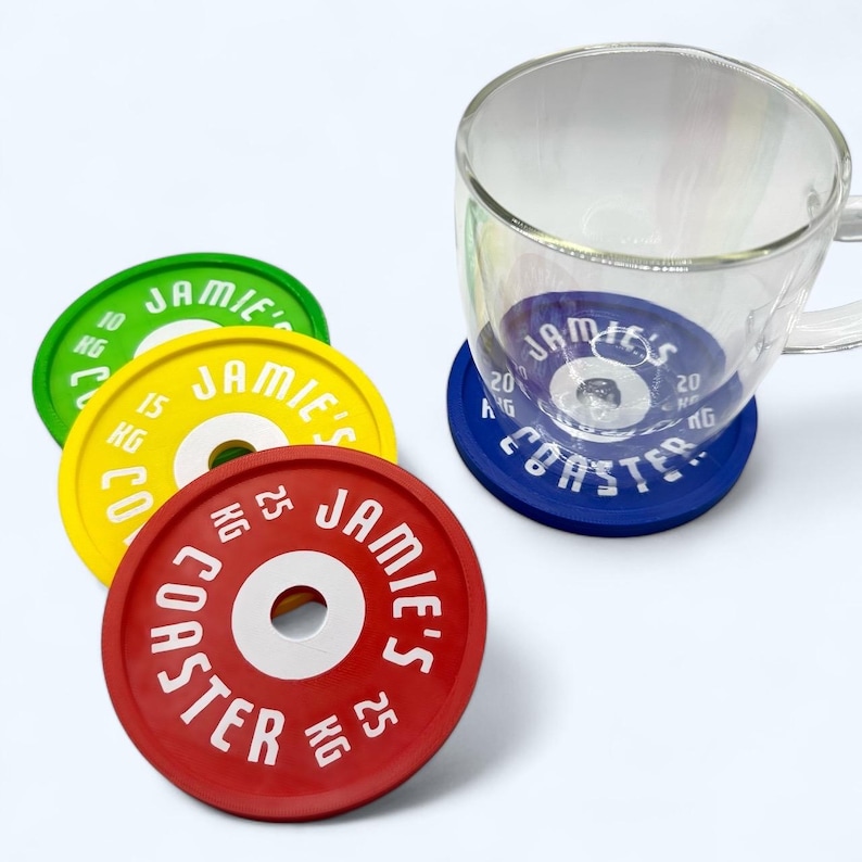 Puede incluir: Un juego de cuatro posavasos circulares con dise&ntilde;o de discos de pesas en verde, amarillo, rojo y azul. Cada posavasos incluye el texto JAMIE'S COASTER, un valor en KG y un orificio central. Una taza de cristal transparente descansa sobre el azul.