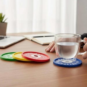 Puede incluir: Un juego de cuatro posavasos circulares dise&ntilde;ados como discos de pesas en verde, amarillo, rojo y azul. El posavasos azul est&aacute; debajo de una taza de cristal transparente con agua. Cada posavasos tiene un orificio central y texto en relieve.