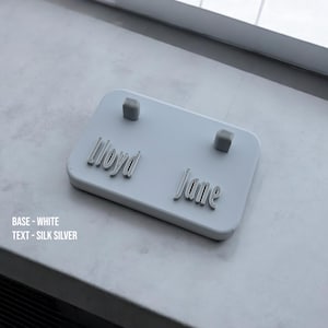 May include: White rectangular nameplate with the names "Lloyd" and "Jane" printed in silver lettering. Two small silver squares are attached to the nameplate.
