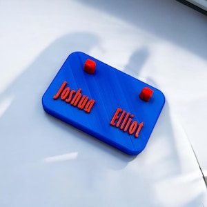 May include: A blue nameplate with the names "Joshua" and "Elliot" printed in red. Two red squares are placed above the names.