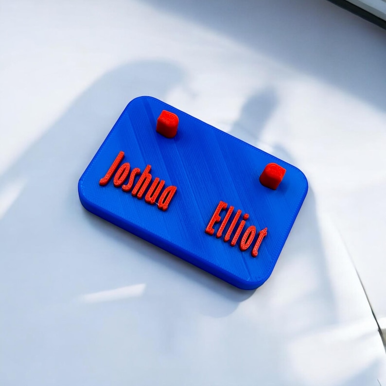 May include: A blue nameplate with the names "Joshua" and "Elliot" printed in red. Two red squares are placed above the names.