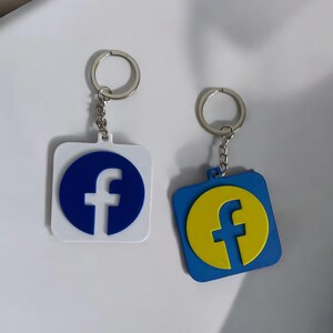 Op de afbeelding: Twee sleutelhangers met een vierkante hanger met het Facebook-logo. De ene sleutelhanger heeft een blauw logo op een witte achtergrond, de andere heeft een geel logo op een blauwe achtergrond.