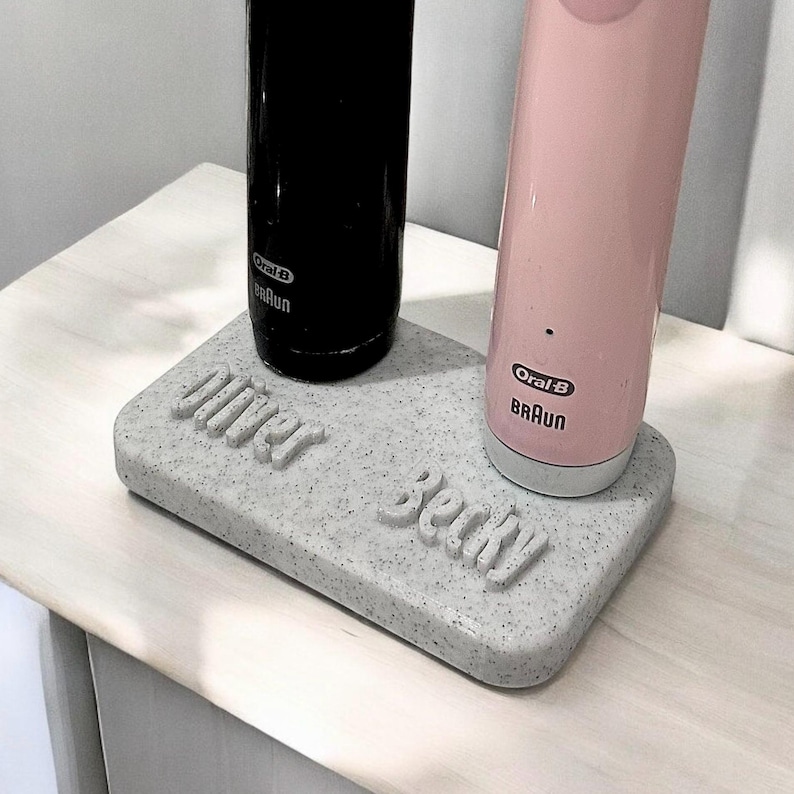 May include: A white, speckled, rectangular charging station with the names "Oliver" and "Becky" written on it. Two electric toothbrushes, one black and one pink, are plugged into the station.