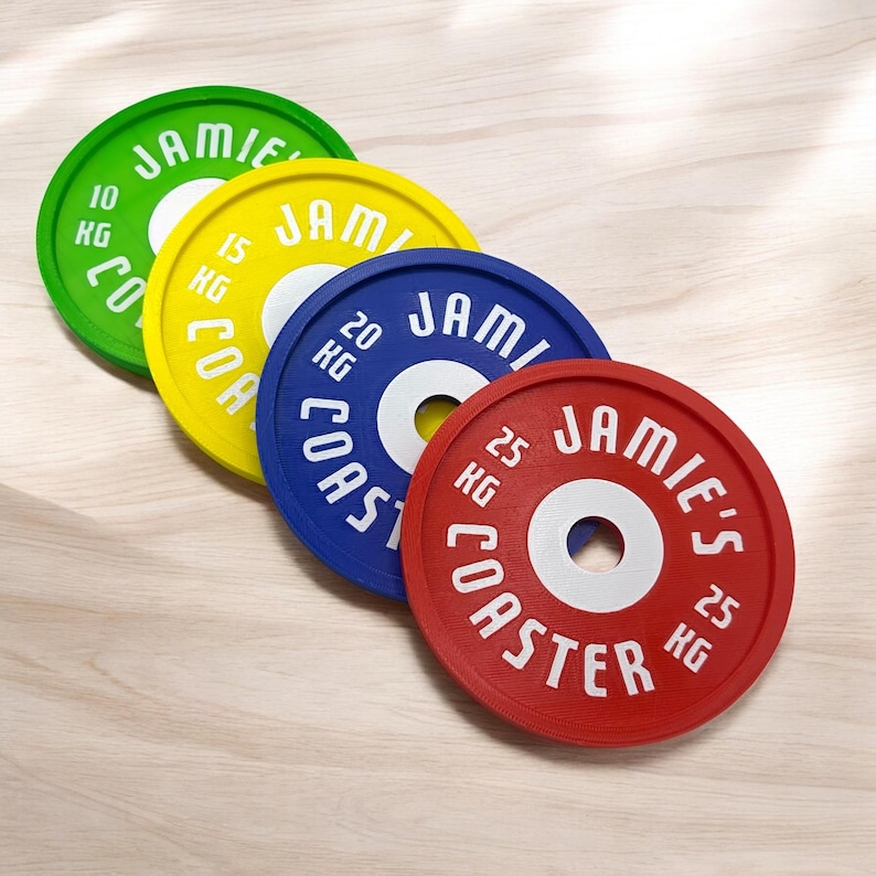 Puede incluir: Cuatro posavasos circulares dise&ntilde;ados como discos de pesas en verde, amarillo, azul y rojo. Cada posavasos presenta el texto JAMIE'S COASTER y marcas de peso de 10 KG, 15 KG, 20 KG y 25 KG, con un orificio central de color blanco.
