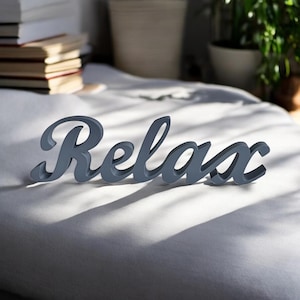 Può includere: Lettere in legno grigie che compongono la parola "Relax" su una superficie bianca.