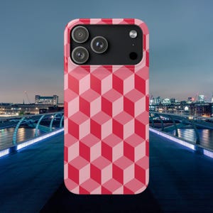 Può includere: Una custodia per telefono rosa con un motivo geometrico a cubo nei toni del rosso. La custodia ha un modulo fotocamera nero con tre lenti e un flash. Lo sfondo mostra un paesaggio urbano con un ponte.