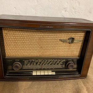Puede incluir: Radio de madera vintage con una cubierta de altavoz tejida y detalles en latón. La radio tiene dos perillas, un dial de sintonización y varios botones blancos. El gabinete es de color marrón oscuro y la radio parece estar en buenas condiciones.