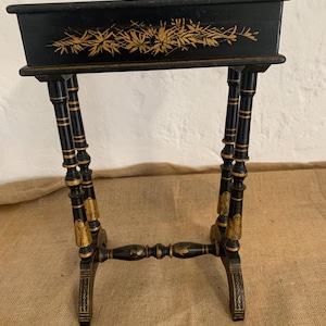 Puede incluir: Soporte rectangular negro y dorado para plantas con diseño floral. El soporte tiene cuatro patas y una parte superior rectangular. Las patas son negras con detalles dorados. La parte superior es negra con un diseño floral dorado.