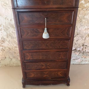 May include: A tall, dark brown wooden chest of drawers featuring six horizontal drawers with a chevron wood grain pattern. Each drawer has a central keyhole, and a light blue decorative tassel hangs from the handle of the second drawer down.