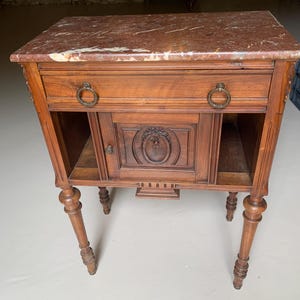 Op de afbeelding: Een houten nachtkastje met een rechthoekig rood marmeren blad, één lade met twee ronde messing ringgrepen en een centrale kastdeur met bloemmotieven. Het meubelstuk staat op vier gedraaide poten en heeft een open onderste plank.