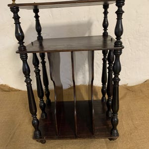 May include: A dark wooden magazine rack with three shelves and decorative turned legs. The top of the rack has small brass accents. The rack is designed to hold magazines and other reading materials. The rack is made of dark wood.