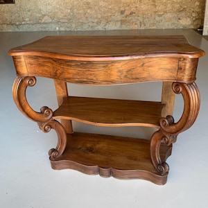Peut inclure: Une console en bois avec un dessus incurvé et deux étagères inférieures. La table présente des supports ornés et enroulés et une finition marron chaud et riche. Ses dimensions sont d'environ 122 cm de large, 46 cm de profondeur et 76 cm de haut.
