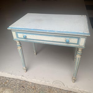 May include: A rectangular wooden table featuring a distressed white finish with light blue painted accents along the edges and drawer front. The table includes a single drawer with two small blue knobs and four slender, fluted legs.