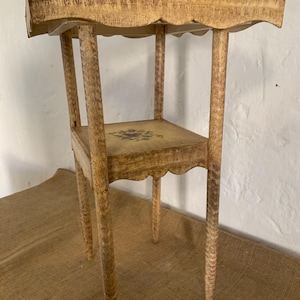 May include: A small, antique, hand-painted side table with a scalloped edge. The table has a distressed, light yellow finish with a floral design on the lower shelf. The legs are slender and tapered, adding to its delicate appearance. The table is approximately 71 cm tall.
