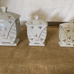 Peut inclure: Trois boîtes de conservation en céramique blanche avec couvercles, ornées de motifs floraux dorés et de détails nervurés horizontaux. Les boîtes portent les inscriptions FARINE, CAFE et THE en lettres dorées sur le devant de chaque récipient.