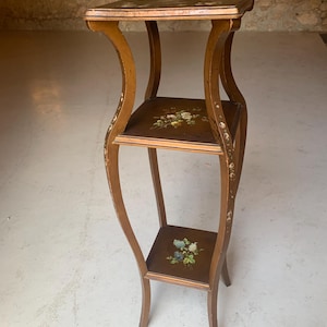Op de afbeelding: Een houten plantenstandaard met drie niveaus, gebogen poten en een donkerbruine afwerking. Elke vierkante plank is voorzien van een handgeschilderd bloemmotief in wit, geel en blauw. De poten zijn versierd met kleine bloemaccenten.