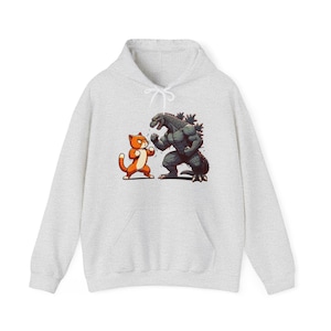 May include: A light grey hoodie with a cartoon illustration of an orange cat and Godzilla in a fighting pose. The cat is orange and white, and Godzilla is grey. The hoodie has a drawstring hood and a front pocket.
