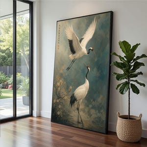 May include: Large framed art print featuring two Japanese cranes. One crane is in flight with outstretched wings, the other standing. The artwork has a muted color palette of blues, whites, and blacks, with a touch of red. The artwork is displayed in a modern interior setting.