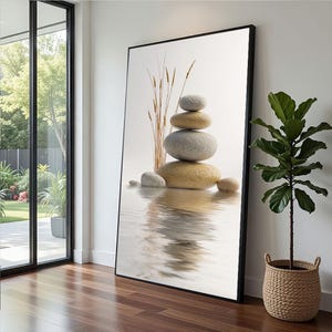 May include: A framed art print featuring a stack of smooth, gray and tan stones, with a few stalks of dried grass. The stones are reflected in the water below, creating a serene and minimalist aesthetic. The artwork is displayed indoors.