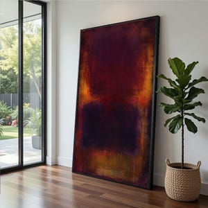 May include: Large abstract painting in a black frame. The artwork features a blend of deep reds, oranges, and purples, creating a textured, color-field effect. The painting leans against a white wall next to a potted plant in a woven basket.