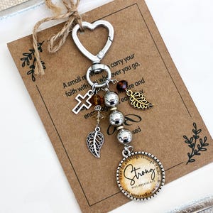 May include: A silver-toned keychain with a heart-shaped clasp, adorned with charms including a cross, leaf, and beads. A circular pendant reads "Strong" with the Bible verse Philippians 4:13. The keychain is attached to a brown card with text.