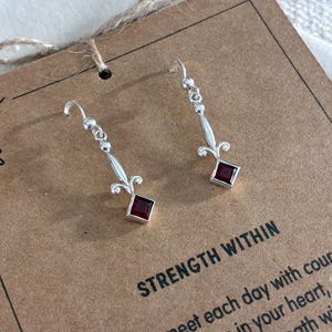 May include: A pair of silver drop earrings featuring a square-cut dark red gemstone at the base of a fleur-de-lis inspired metal design. The earrings are attached to a brown card printed with the text STRENGTH WITHIN.