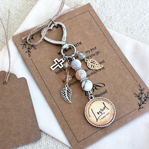 May include: A silver-toned heart-shaped keychain featuring a cross charm, two leaf charms, and three marbled beads. A circular pendant at the bottom displays the text "Loved 1 JOHN 4:19" in black script on a tan background, attached to a card.