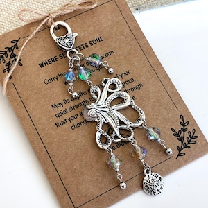 May include: A silver-toned octopus charm with dangling iridescent beads and a sand dollar pendant. The charm is attached to a clasp and tied to a brown card with text that reads "WHERE SEETS SOUL". The background is white.
