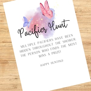 May include: A white paper with the text "Pacifier Hunt" in black script, decorated with watercolor butterflies in pink and blue. The text explains the game rules for a baby shower. The background is a wooden surface.