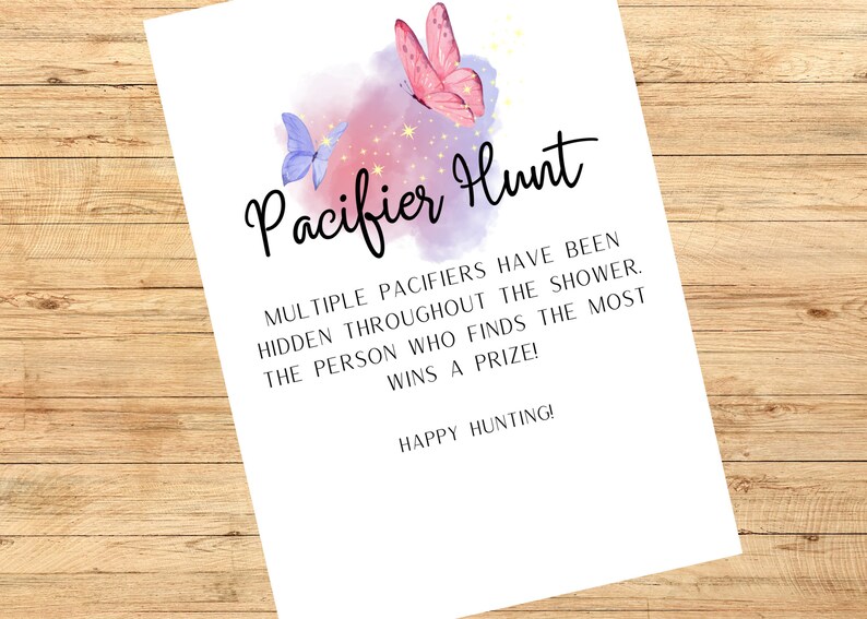 May include: A white paper with the text "Pacifier Hunt" in black script, decorated with watercolor butterflies in pink and blue. The text explains the game rules for a baby shower. The background is a wooden surface.