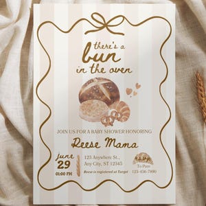 May include: Baby shower invitation with a beige and white striped background. The invitation features a brown decorative border and text that reads "there's a bun in the oven." Watercolor illustrations of bread and pastries are included.