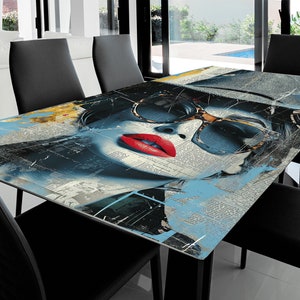 Può includere: Tavolo da pranzo rettangolare con un design artistico sorprendente. La superficie del tavolo presenta il volto di una donna con occhiali da sole e labbra rosse, su uno sfondo di testo e schizzi di vernice blu e gialla. Sedie nere.