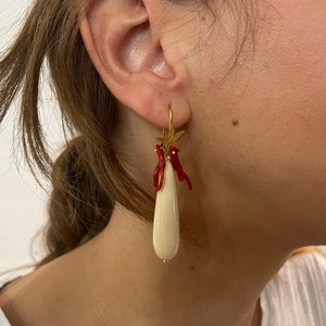 Op de afbeelding: Goudkleurige oorbellen met accenten van zeesterren en rood koraal. Elke oorbel heeft een lange, druppelvormige, crèmekleurige hanger. De oorbellen zijn ontworpen om te bungelen en het licht te vangen.
