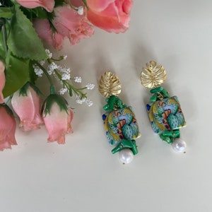 May include: A pair of gold and green earrings with a floral design and a pearl accent. The earrings feature a green and blue tile-like design with gold accents.
