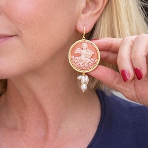 May include: A round earring featuring a peach-colored cameo of a winged cherub playing a musical instrument, set within a gold-toned beaded frame. Three small, irregular white pearls hang from the bottom of the circular pendant on a gold hook.