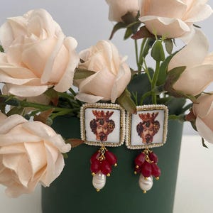 Puede incluir: Un par de pendientes de diseño cuadrado con un retrato de un hombre con corona y barba. Los pendientes están adornados con cuentas rojas y una perla colgante.