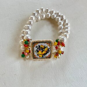 Puede incluir: Un brazalete de perlas blancas con un dije cuadrado de cerámica con un diseño de limón colorido. El dije está rodeado de pequeñas cuentas rojas, verdes y amarillas.