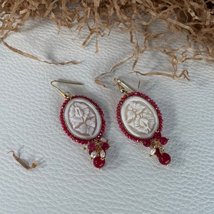 May include: A pair of white cameo earrings with a floral design, surrounded by red beads and accented with white pearls.