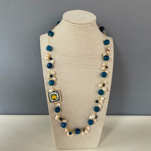 Puede incluir: Un collar largo con cuentas azules y blancas y una cadena dorada. El collar presenta un pequeño colgante cuadrado con un diseño floral.