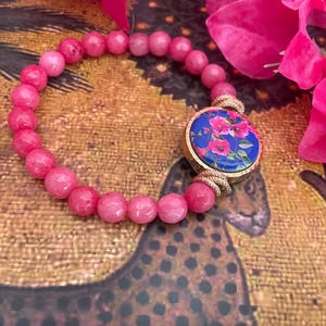 Könnte beinhalten: Rosa Perlenarmband mit einem goldfarbenen, kreisförmigen Anhänger mit floralem Design auf blauem Hintergrund. Das Armband besteht aus kleinen, runden rosa Perlen. Der Anhänger ist mit goldfarbenen Spiralen akzentuiert. Rosa Blumen sind im Hintergrund.