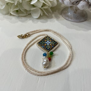 Puede incluir: Un collar de perlas blancas con un colgante cuadrado con un diseño floral azul y dorado. El colgante está acentuado con un grupo de piedras preciosas de colores y una perla grande.