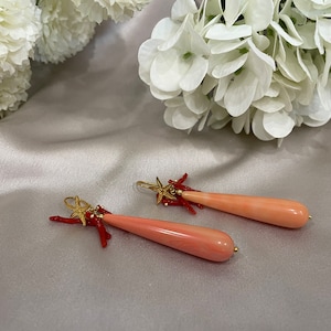May include: A pair of coral earrings with gold accents and red coral branches. The earrings are teardrop shaped and have a pink coral color.