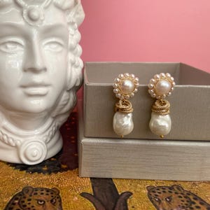 May include: A pair of pearl drop earrings. Each earring has a large, irregular white pearl hanging below a cluster of smaller pearls and gold-coloured metal details. The earrings are presented on a neutral surface against a pink background.