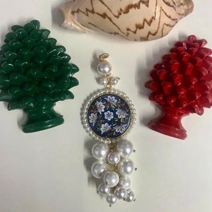 May include: A gold pendant with a navy blue floral design, encircled by faux pearls, and a cluster of white faux pearls. Beside it are green and red pinecone ornaments and a seashell.
