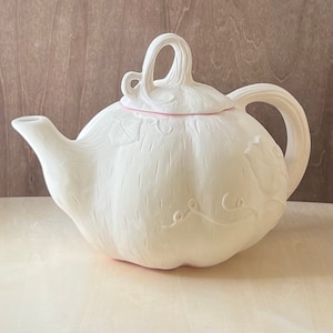 Puede incluir: Tetera de cerámica blanca con forma de calabaza y un diseño de vid detallado. La tetera tiene una tapa con un asa que se asemeja a un tallo de calabaza.