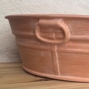 Puede incluir: Una jardinera ovalada de color terracota con una superficie texturizada y un asa. La jardinera tiene un borde redondeado y está sobre una superficie de madera. El asa es un diseño de bucle.