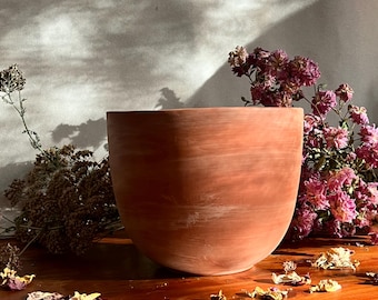 May include: A terracotta-colored ceramic bowl with a smooth, rounded shape. The bowl is sitting on a wooden surface with dried flowers and petals scattered around it.